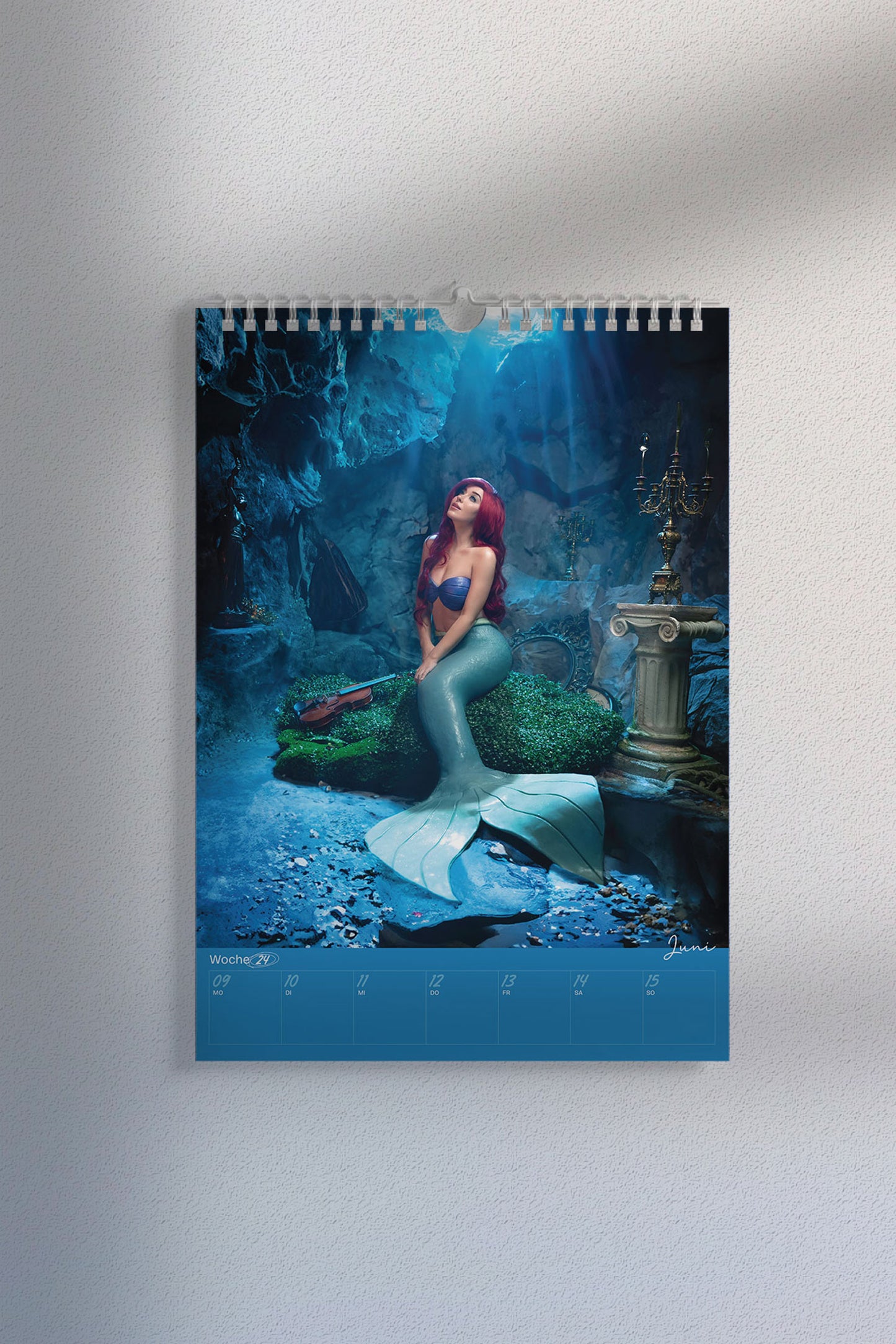 Kalenderseite mit Cosplay-Foto einer Meerjungfrau mit roter Perücke, türkisfarbenem Fischschwanz und Muscheloberteil. Szene spielt in einer Unterwasser-Fantasy-Kulisse. Unten der Wochenplaner „Juni, Woche 24“.