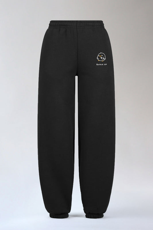 Schwarze Jogginghose mit elastischem Bund und Bündchen am Beinabschluss. Auf dem linken Oberschenkel ist ein kleines Motiv mit einer weißen, rund gezeichneten Ente mit orangefarbenem Schnabel und Füßen aufgestickt. Darunter steht in Großbuchstaben der Schriftzug „QUACK UP“.
