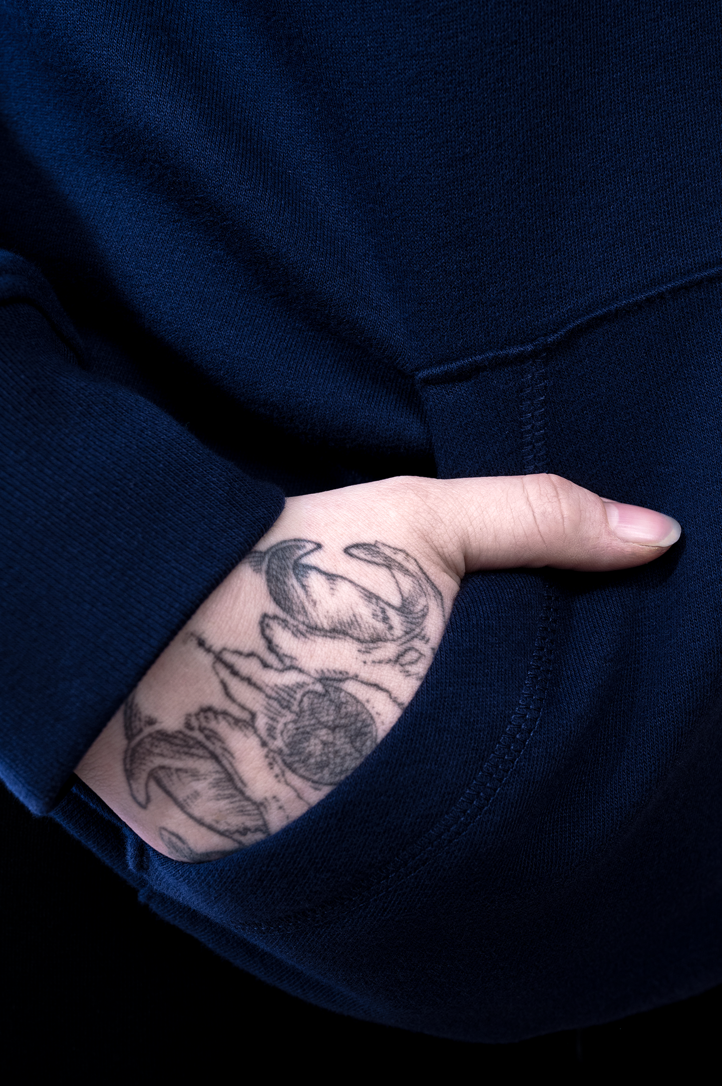 Detailaufnahme der Kängurutasche des Hoodies mit tätowierter Hand in der Tasche, Fokus auf Stoffstruktur und Nahtverarbeitung.