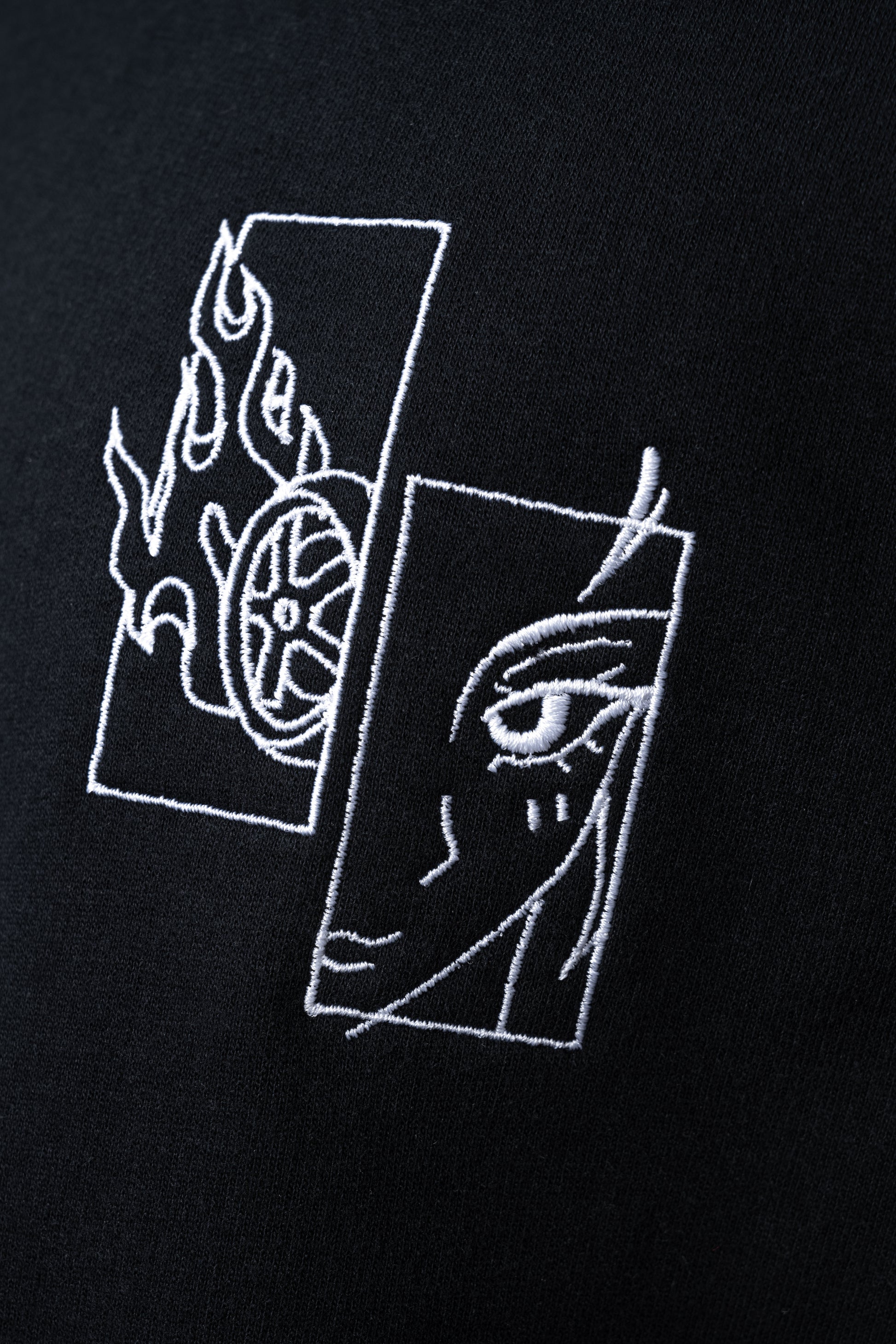 Nahaufnahme des weißen Stickmotivs: linkes Rechteck zeigt ein Flammenrad, rechtes ein stilisiertes Gesicht mit geschlossenem Auge.