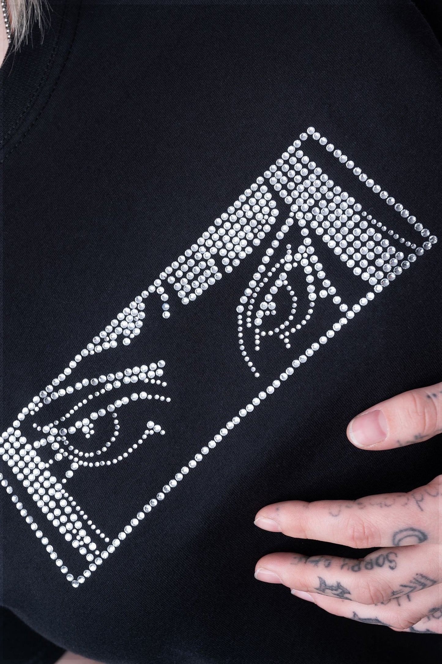 Nahaufnahme eines schwarzen T-Shirts mit rechteckigem Strassmotiv auf der Brust. Das Design besteht aus zwei stilisierten Augen, die mit zahlreichen kleinen, silberfarbenen Strasssteinen umgesetzt sind. Eine tätowierte Hand berührt den Stoff von unten.