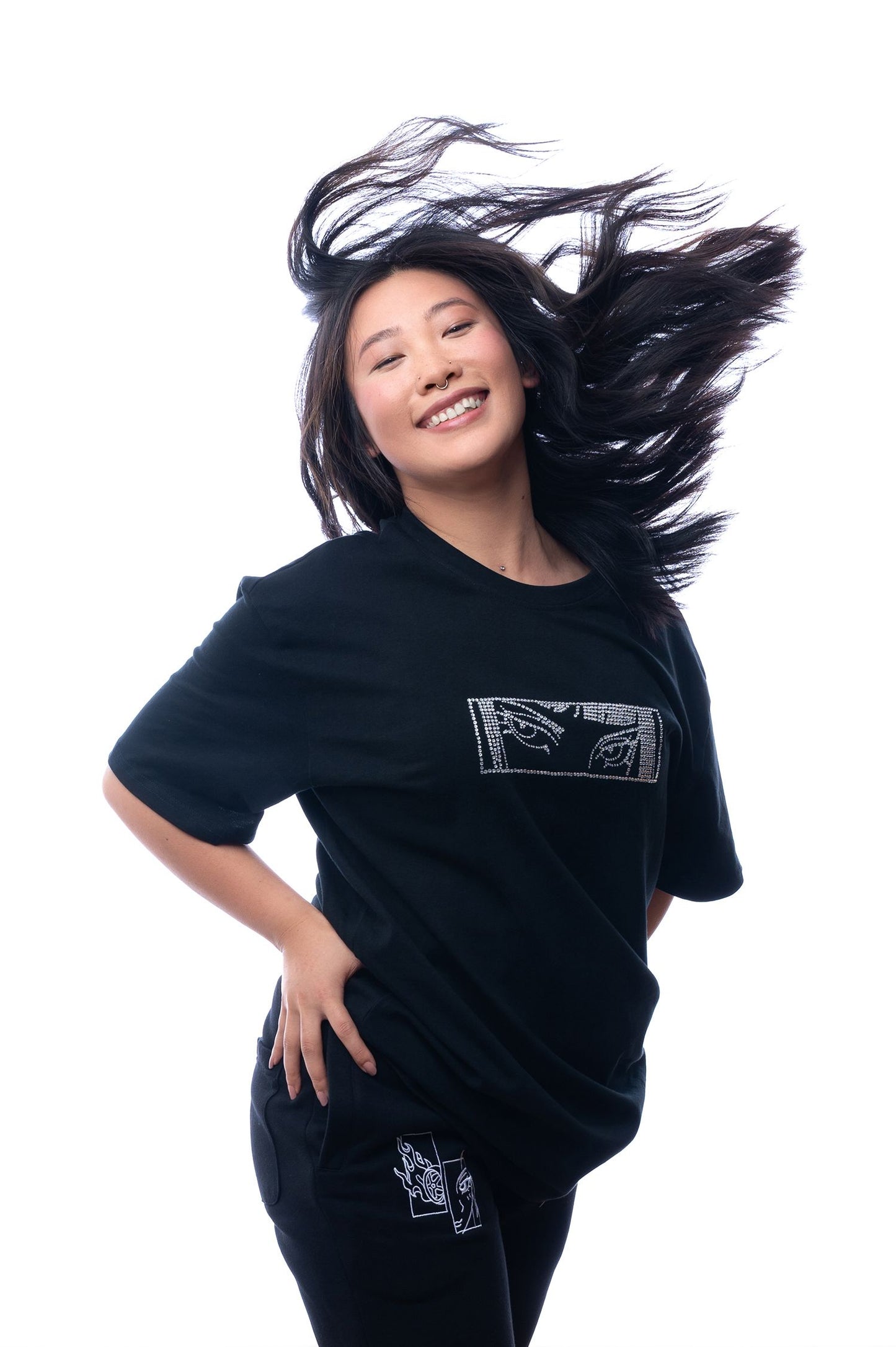 Schwarzes Oversize-T-Shirt mit glitzerndem Brustmotiv, bestehend aus zwei stilisierten Augen in einer rechteckigen Fläche aus silberfarbenen Strasssteinen. Das T-Shirt wird locker getragen, die Pose ist frontal und der Hintergrund zeigt intensive rote Lichtsäulen mit Nebeleffekt.