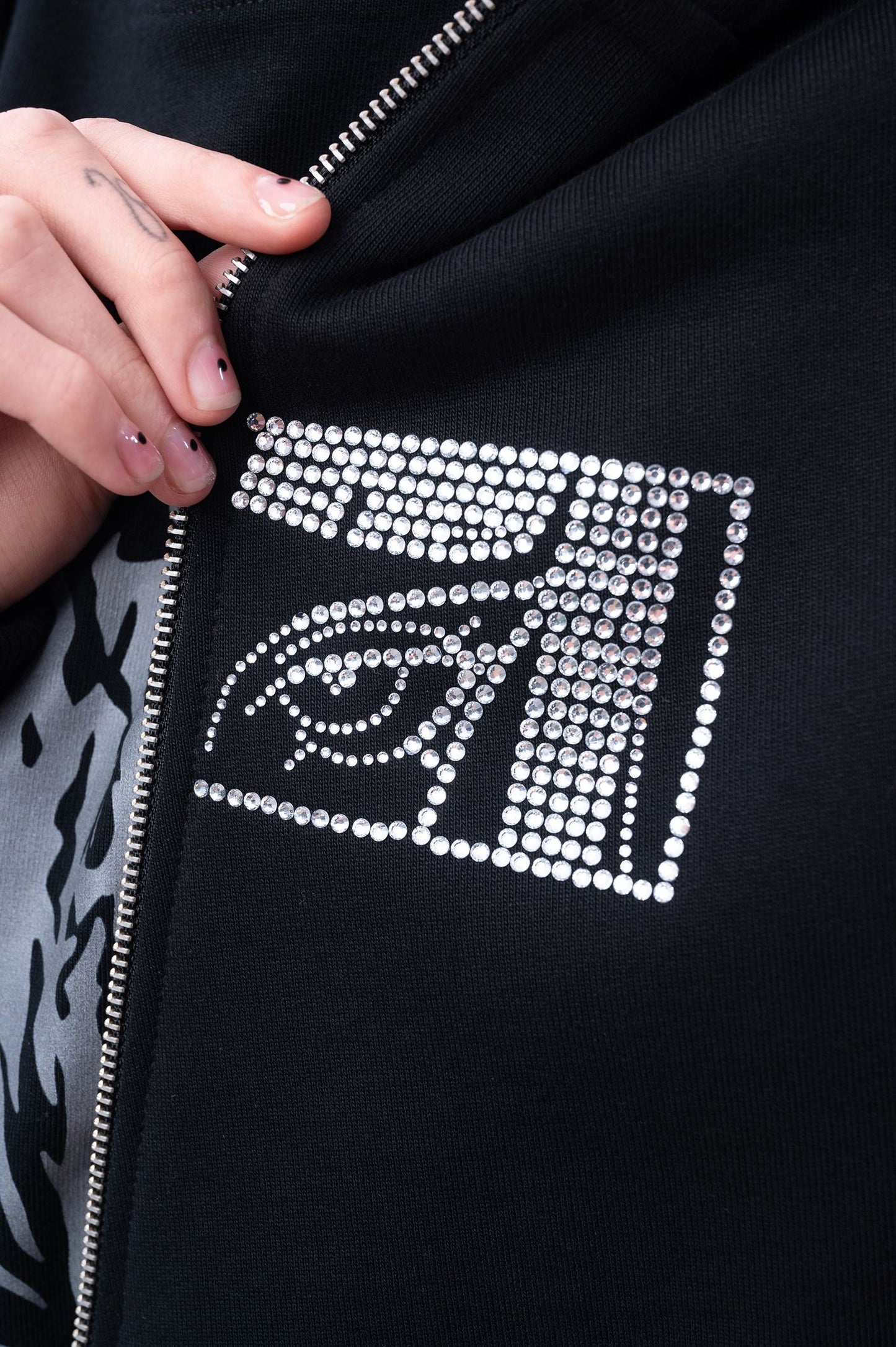 Nahaufnahme des linken Brustbereichs eines schwarzen Zip-Hoodies mit silbernem Reißverschluss und funkelndem Strassmuster in Form eines stilisierten Auges im rechteckigen Rahmen.