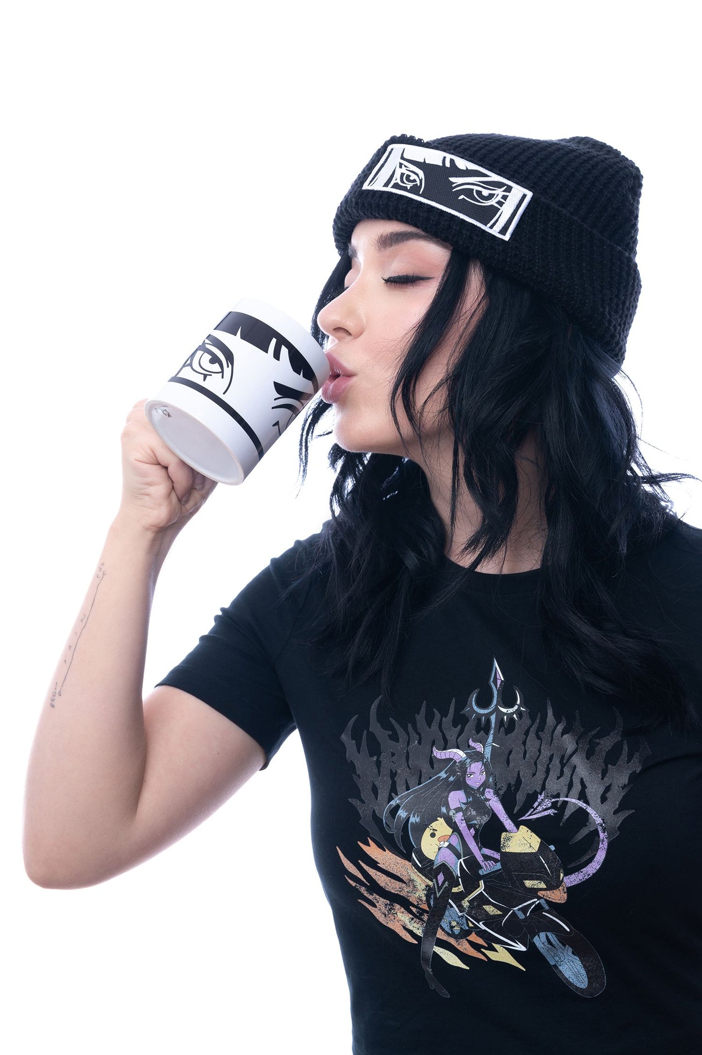 Weiße Tasse mit schwarzem Augenmotiv wird zum Mund geführt. Die trinkende Person trägt ein schwarzes Beanie mit identischem Augenmotiv auf dem Aufnäher