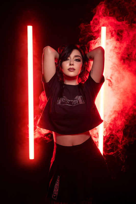 Schwarzes, kurz geschnittenes T-Shirt mit glitzerndem Strassmotiv auf der Brust. Das Design zeigt zwei stilisierte Augen in einem rechteckigen Rahmen, bestehend aus kleinen, funkelnden Steinen. Das Shirt wird in roter Lichtszene mit aufsteigendem Nebel präsentiert.