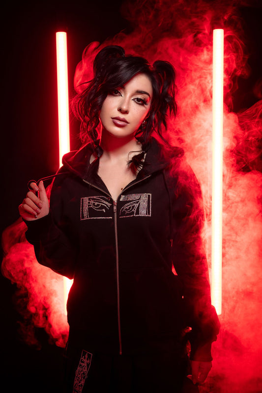 Model mit schwarzem Zip-Hoodie vor rotem Licht und Nebel. Der Hoodie zeigt zwei spiegelverkehrte Augenmotive aus Strasssteinchen auf Brusthöhe, mittig geteilt durch einen silbernen Reißverschluss. Das Model hält eine Kordel der Kapuze locker in der Hand.