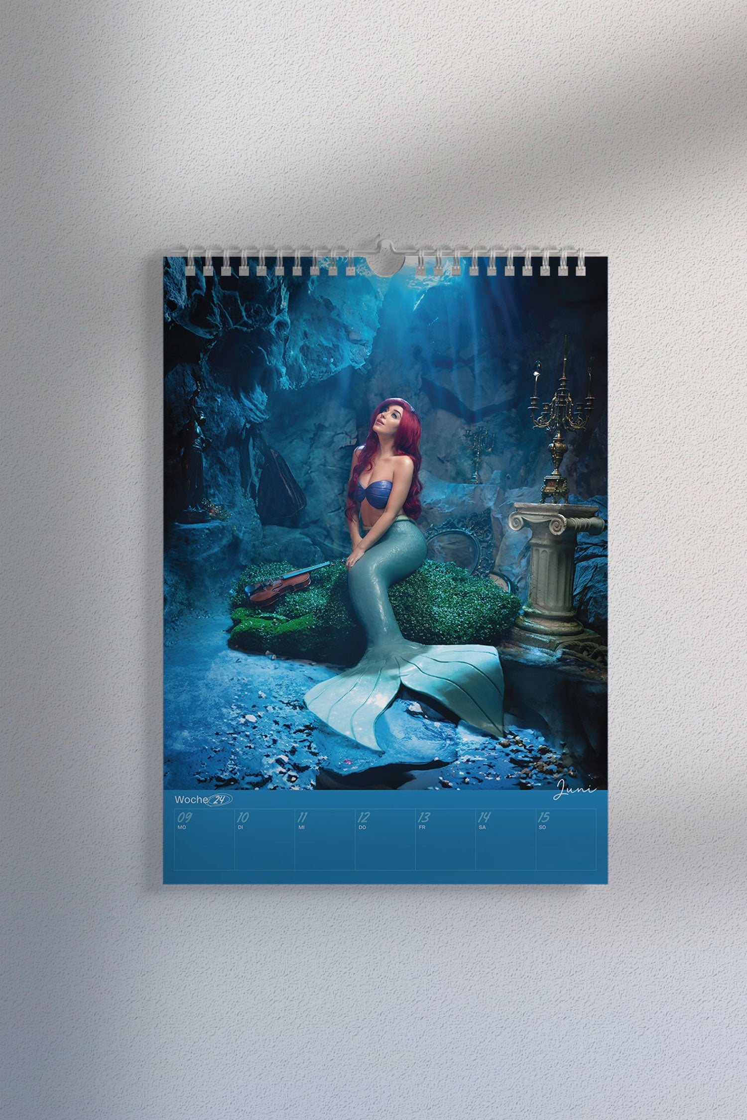 Kalenderseite mit Cosplay-Foto einer Meerjungfrau mit roter Perücke, türkisfarbenem Fischschwanz und Muscheloberteil. Szene spielt in einer Unterwasser-Fantasy-Kulisse. Unten der Wochenplaner „Juni, Woche 24“.