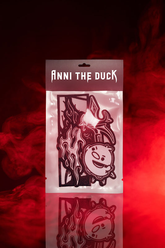 Set aus drei gestickten Patches in transparenter Verpackung mit schwarzer Kopfkarte und weißem Schriftzug „Anni the Duck“. Die Patches zeigen eine stilisierte Flamme mit Diamant, ein grafisches Auge im Rechteck und ein rundes Cartoon-Gesicht mit Grinsen. Präsentiert vor rotem Rauchhintergrund.