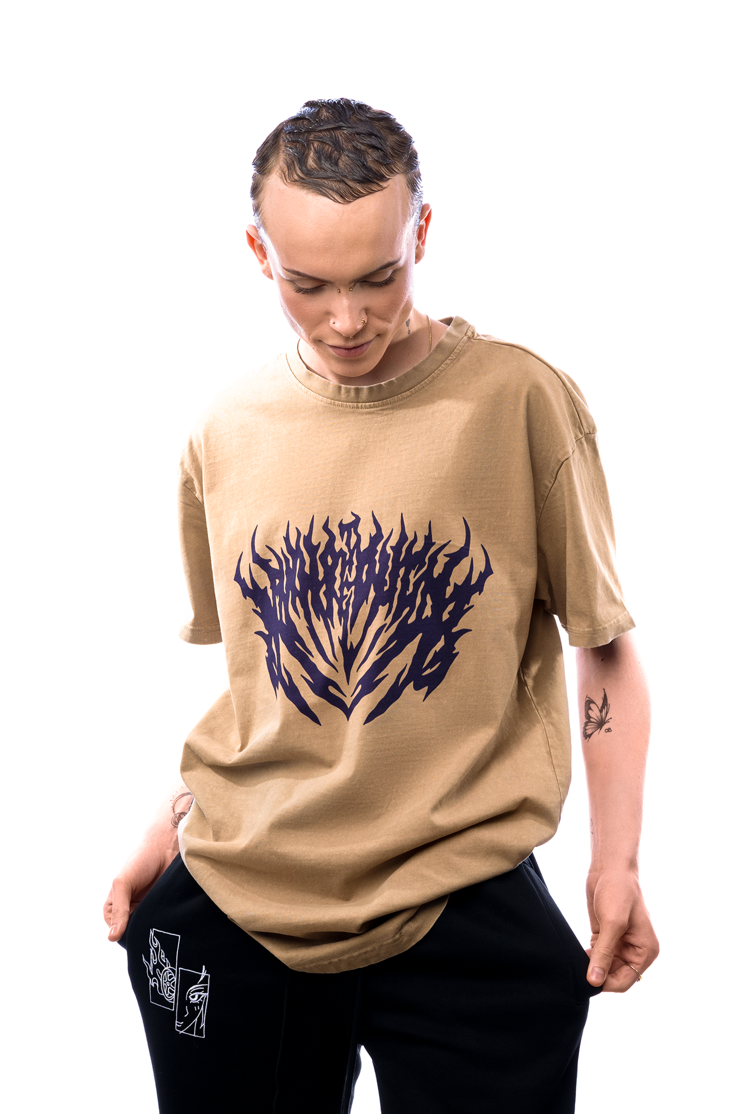 Locker geschnittenes braunes T-Shirt mit violettem Flammenprint, getragen von einer Person mit kurzen Haaren, die nach unten blickt.