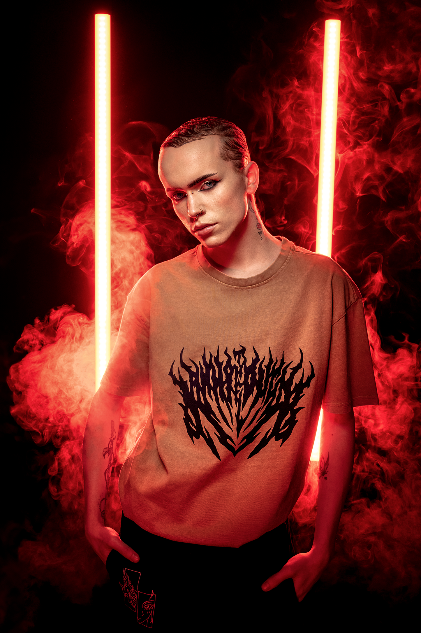 	Braunes Shirt mit schwarzem Flammenschriftzug, getragen von einer Person mit intensiver Schminke vor rotem Nebel und Leuchtröhren.