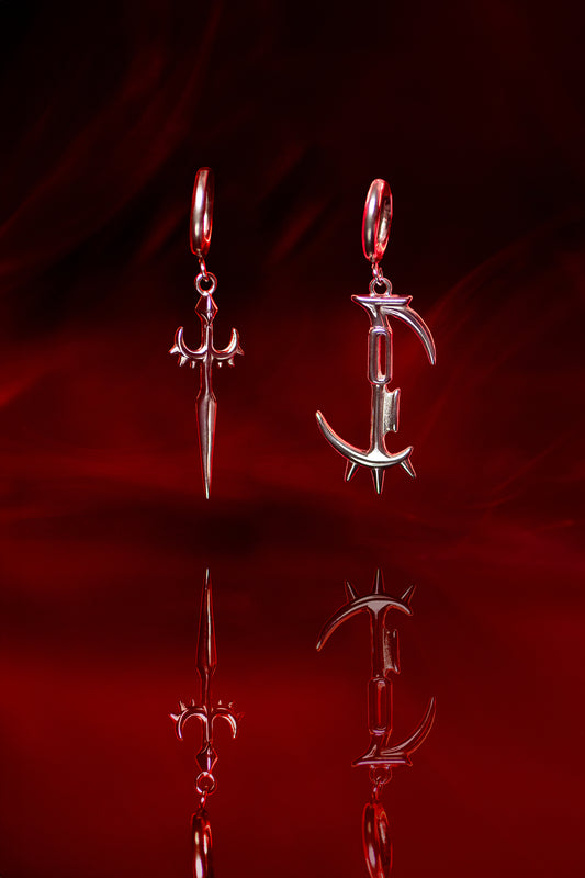 Paar silberner Ohranhänger, links ein Dolchdesign, rechts ein grafischer „C“-Anhänger mit Dornen. Die Schmuckstücke hängen nebeneinander vor rotem Hintergrund und spiegeln sich im glänzenden Untergrund.