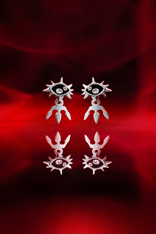Paaraufnahme zweier silberner Ohrstecker in Form eines Auges mit Strahlenkranz und dreizackigem Anhänger, gespiegelt auf glänzend rotem Untergrund.