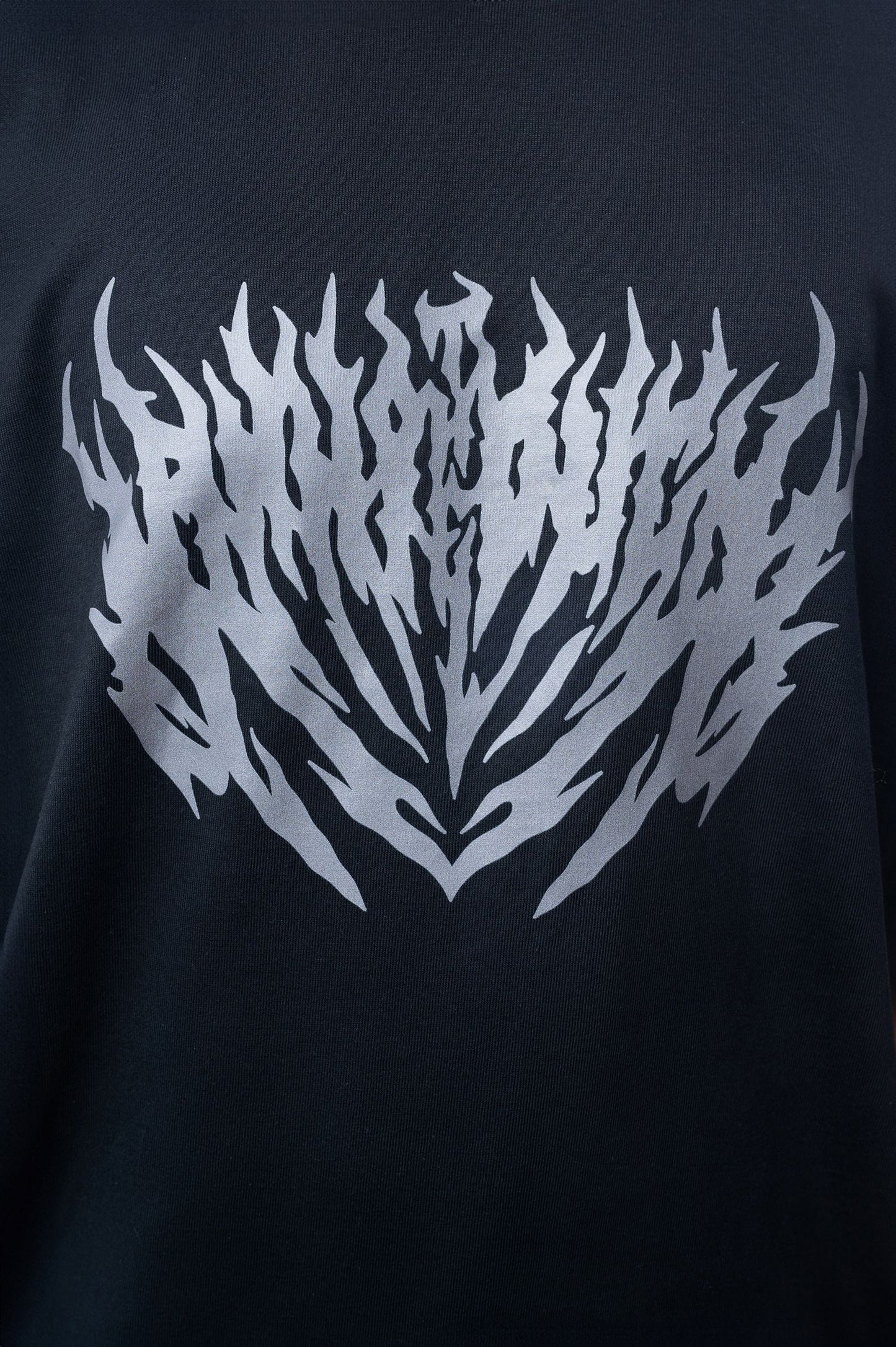 Nahaufnahme eines schwarzen T-Shirts mit großem silbergrauen Print auf der Brust. Der Druck zeigt ein symmetrisch ausgerichtetes, stilisiertes Flammenmuster mit spitz zulaufenden Formen.