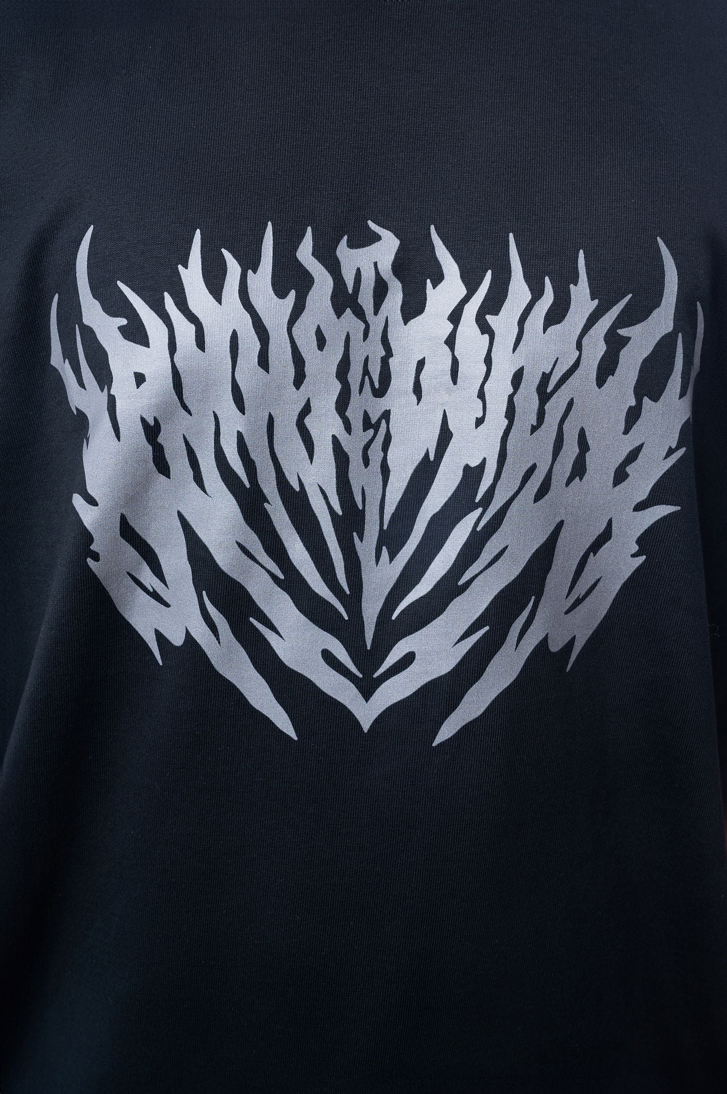 Nahaufnahme eines schwarzen T-Shirts mit großem silbergrauen Print auf der Brust. Der Druck zeigt ein symmetrisch ausgerichtetes, stilisiertes Flammenmuster mit spitz zulaufenden Formen.