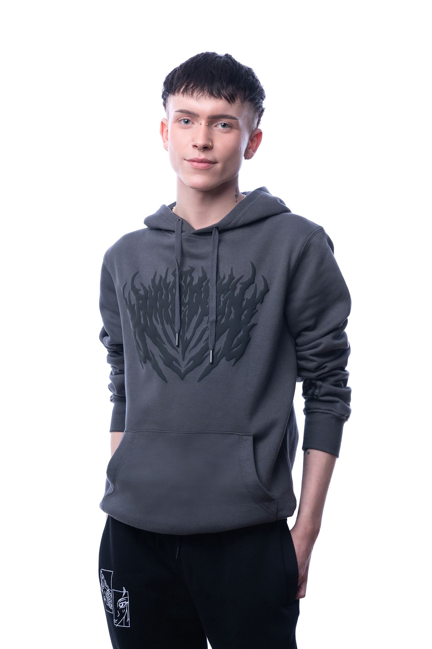 Dunkelgrauer Kapuzenpullover mit mittigem, großflächigem schwarzen Flammenprint auf der Brust. Der Hoodie hat eine Kängurutasche und breite Kordeln mit Metallabschlüssen. Getragen von einer Person in neutraler Studioaufnahme.