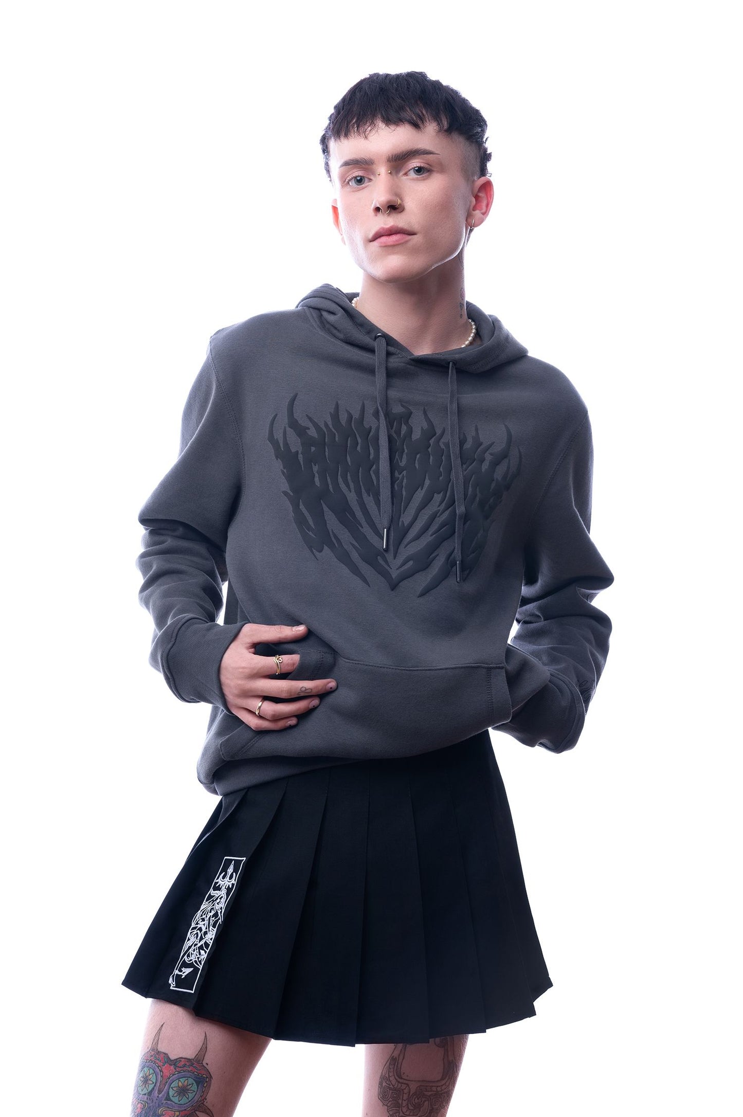 Dunkelgrauer Hoodie mit schwarzem Flammenprint auf der Vorderseite. Die Person trägt dazu einen schwarzen Faltenrock mit weißem grafischen Print auf dem linken Bein. Studioaufnahme mit Blick auf das ganze Oberteil.