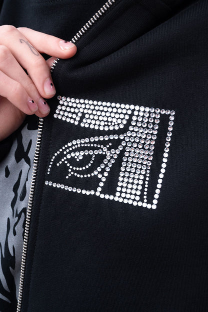 Nahaufnahme des linken Brustbereichs eines schwarzen Zip-Hoodies mit silbernem Reißverschluss und funkelndem Strassmuster in Form eines stilisierten Auges im rechteckigen Rahmen.