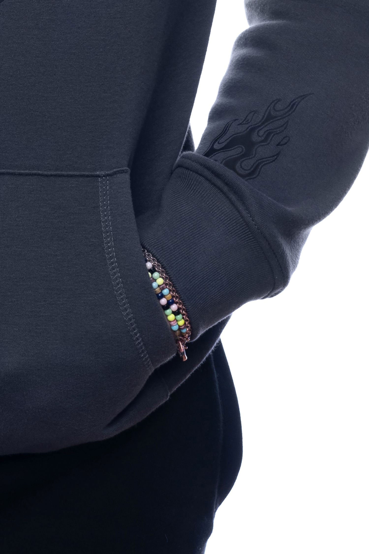 Nahaufnahme des rechten Ärmels des dunkelgrauen Hoodies. Sichtbar ist ein schwarzes Flammenmotiv nahe der Naht sowie eine Hand mit buntem Perlenarmband in der Bauchtasche.