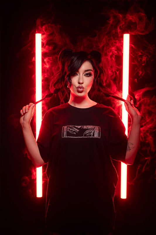 Schwarzes Oversize-T-Shirt mit glitzerndem Brustmotiv, bestehend aus zwei stilisierten Augen in einer rechteckigen Fläche aus silberfarbenen Strasssteinen. Das T-Shirt wird locker getragen, die Pose ist frontal und der Hintergrund zeigt intensive rote Lichtsäulen mit Nebeleffekt.