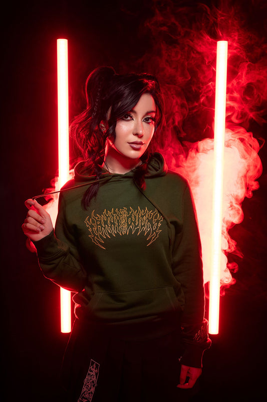 Grüner Kapuzenpullover mit orangefarbener Frontgrafik, getragen in rotem Lichtsetting mit Rauch, kombiniert mit schwarzem Faltenrock.