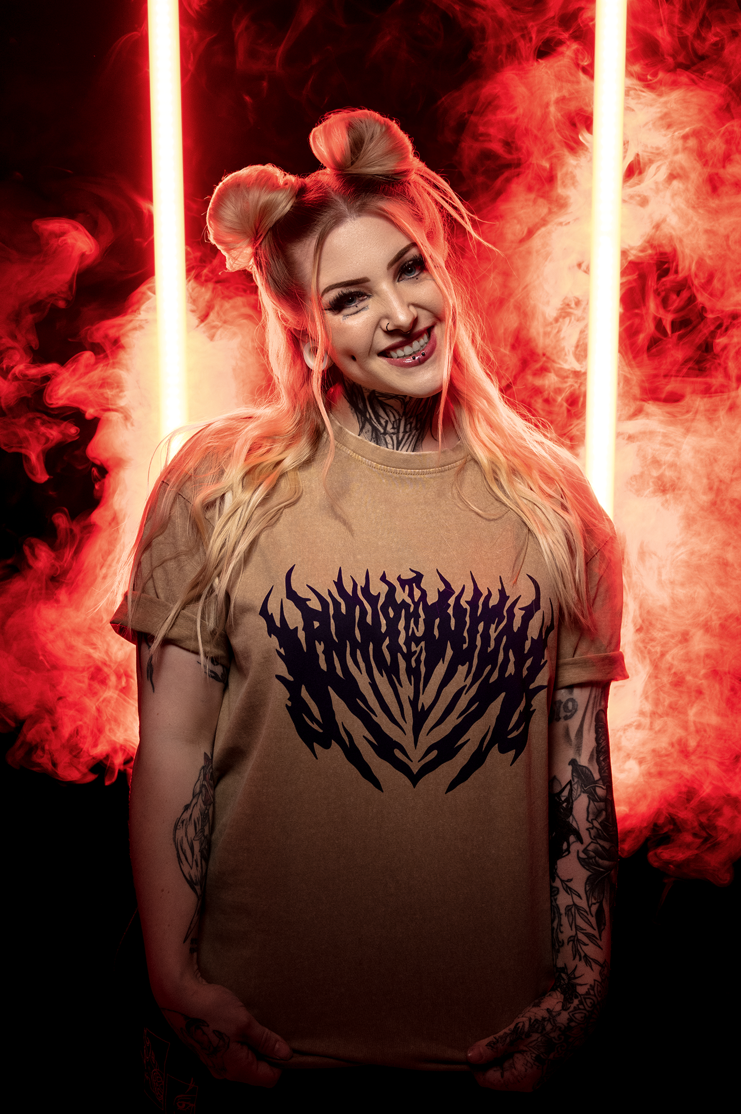 Braun-beiges Shirt mit großem, schwarzem Flammen-Schriftzug, getragen von einer blondhaarigen Person mit Duttfrisur vor rot vernebelter Szenerie.