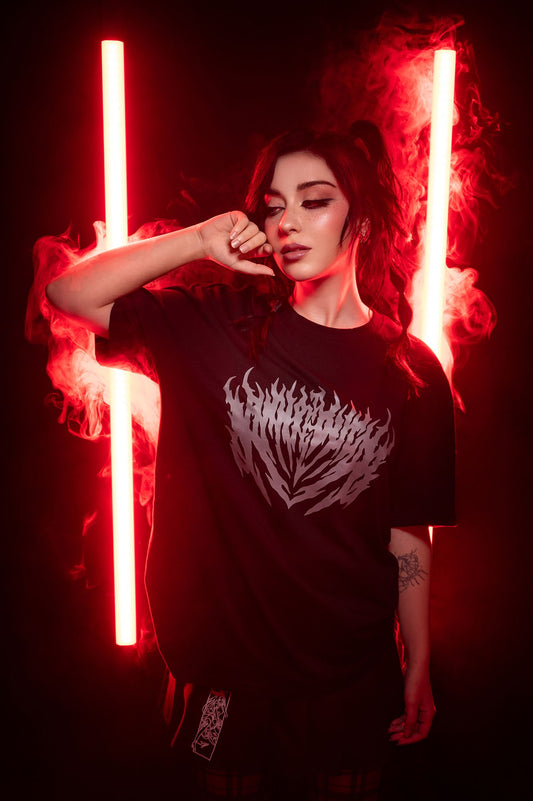 Schwarzes T-Shirt mit reflektierendem silbergrauem Frontprint in flammenartiger Symmetrie. Das Shirt wird von einer Person mit Zöpfen in roter Lichtszene getragen, der Druck reflektiert das Umgebungslicht deutlich sichtbar.