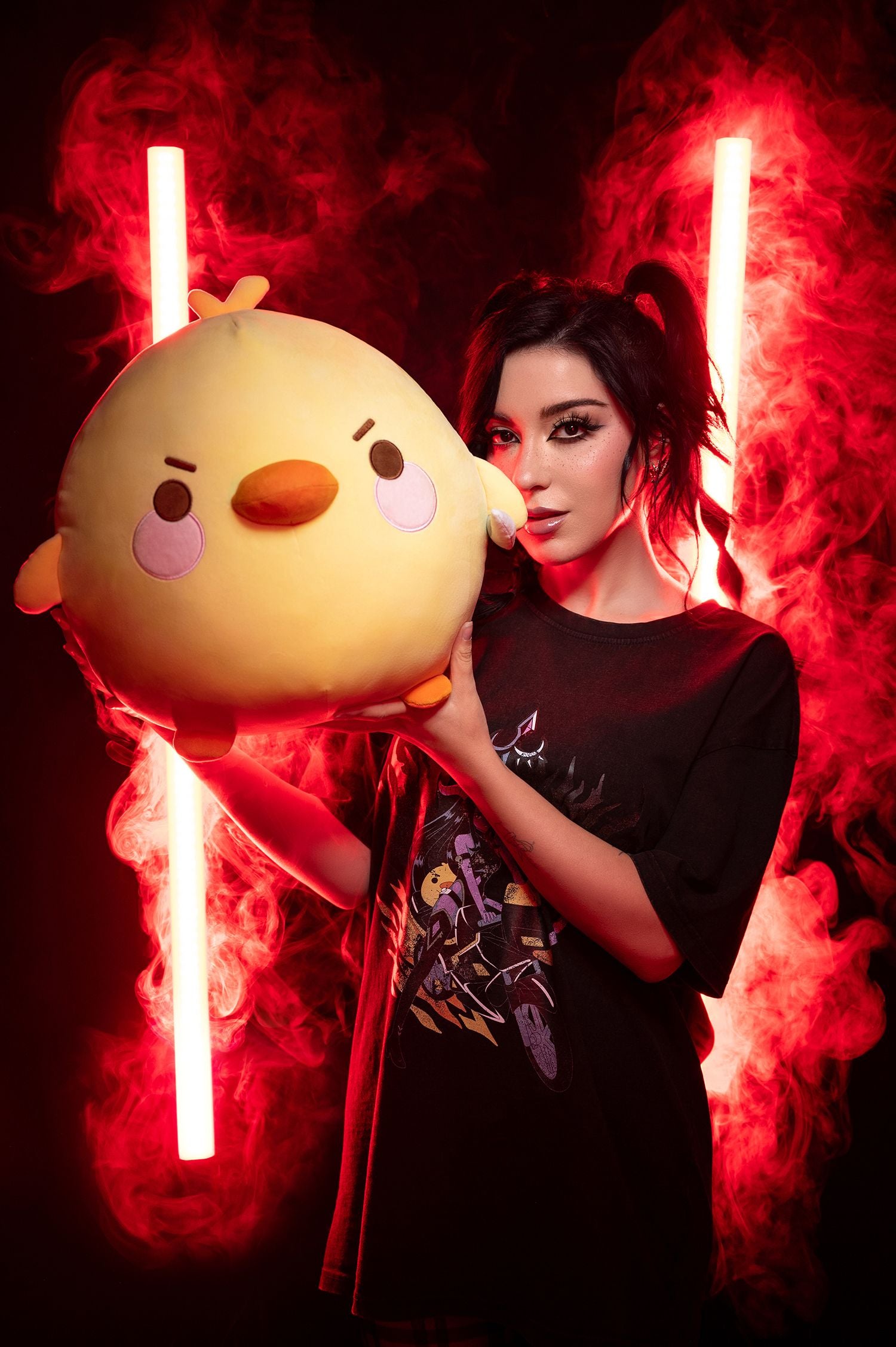 Person in schwarzem Shirt mit Aufdruck posiert vor rotem Nebellicht mit einem großen Enten-Plüschtier in der Hand. Die Ente ist gelb mit rosa Wangen, orangefarbenem Schnabel und entschlossenem Blick.
