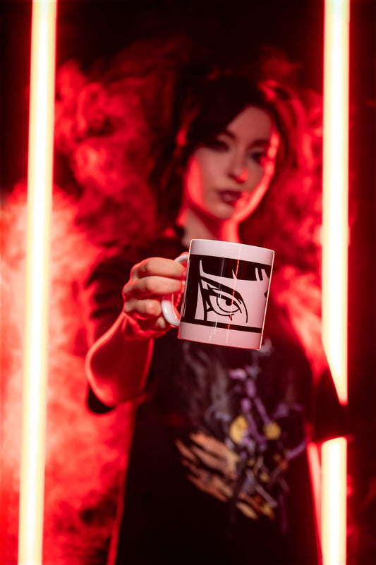 Weiße Tasse mit schwarzem, stilisiertem Augenmotiv wird frontal zur Kamera gehalten. Die Szene ist in rotem Licht mit Rauchinszenierung aufgenommen.