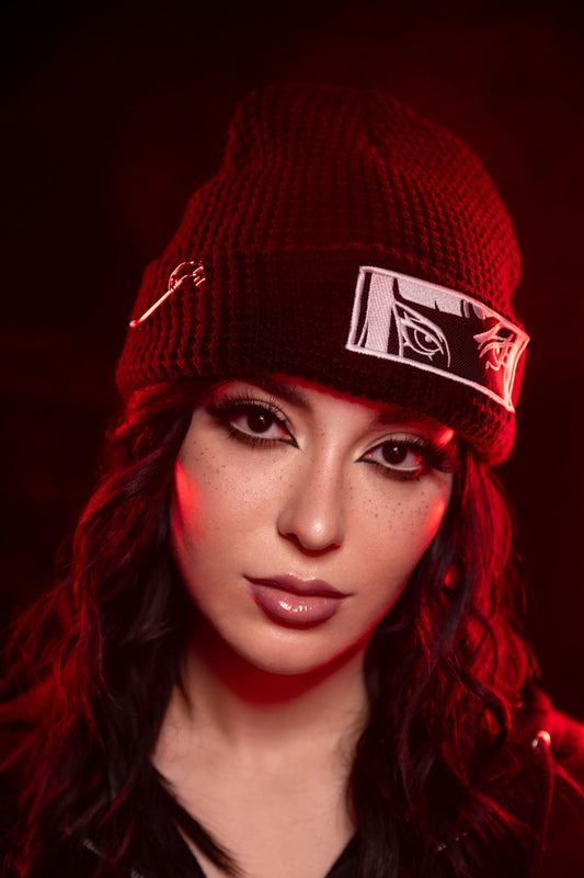 Nahaufnahme einer schwarzen Beanie mit grobem Strickmuster, getragen von einer Person mit langen schwarzen Haaren. Auf dem umgeschlagenen Saum ist ein rechteckiger Patch mit weißer Augenstickerei im Comicstil zu sehen. Links ist eine silberfarbene Sicherheitsnadel befestigt. Rotes Licht umrahmt die Szene.