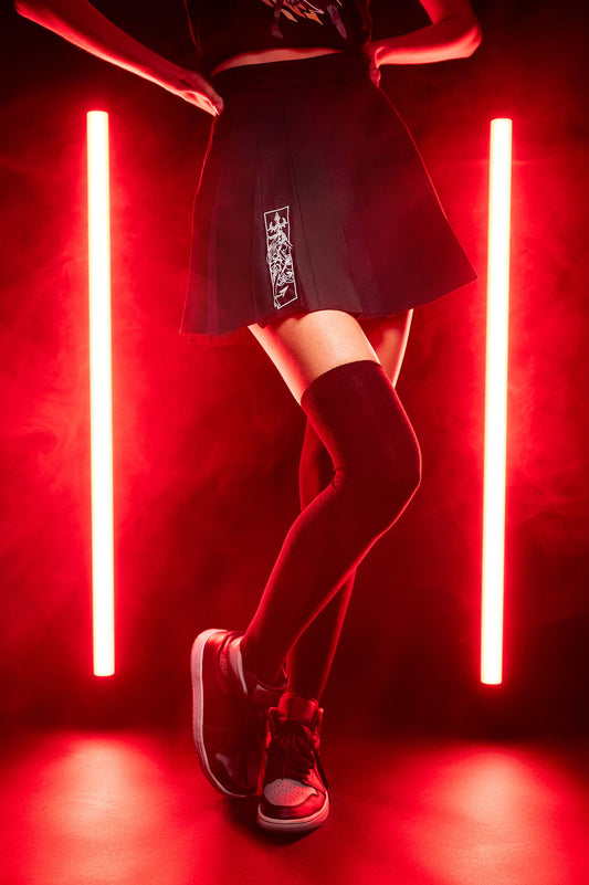 Schwarzer Faltenrock mit weißem Stickmotiv auf der Vorderseite. Das Bild ist in rotem Licht fotografiert, die Person trägt schwarze Kniestrümpfe und Sneakers, steht mit überkreuzten Beinen.
