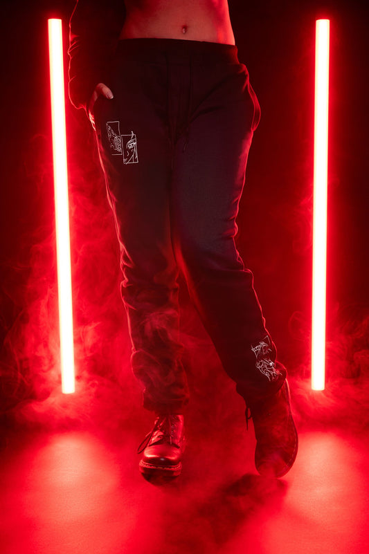 Schwarze Jogginghose mit weißen Stickmotiven auf beiden Beinen, getragen in Szene mit roten Neonlichtern und Bodennebel. Linkes Bein zeigt zwei rechteckige grafische Illustrationen auf Oberschenkelhöhe, rechtes Bein eine komplexe Liniengrafik am Unterschenkel.